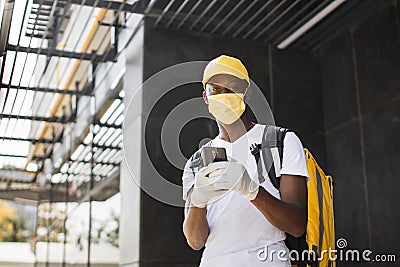 African American male courier food delivery in face mask with yellow thermal backpack Stock Photo