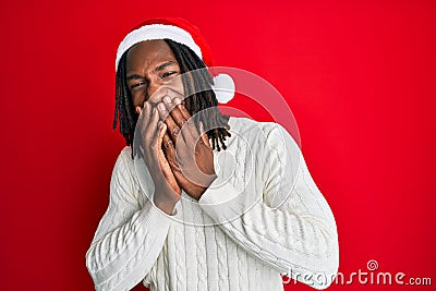 African american man with braids wearing christmas hat laughing and embarrassed giggle covering mouth with hands, gossip and Stock Photo