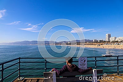 A African American man sitting on a bench on a wooden pier with vast blue ocean water, hotels and office buildings the cityscape Editorial Stock Photo
