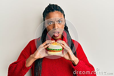African american woman with braids eating hamburger in shock face, looking skeptical and sarcastic, surprised with open mouth Stock Photo