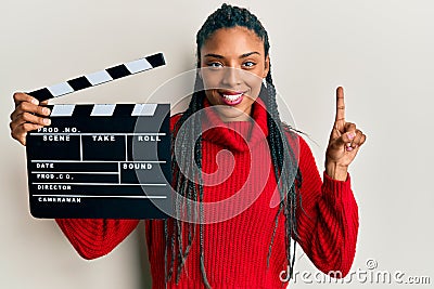 African american woman with braids holding video film clapboard smiling with an idea or question pointing finger with happy face, Stock Photo