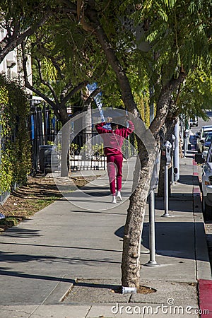 An African American woman with long hair wearing a red sweat suit walking along a sidewalk lined with lush green trees Editorial Stock Photo