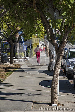 An African American woman with long hair wearing a red sweat suit walking along a sidewalk lined with lush green trees Editorial Stock Photo