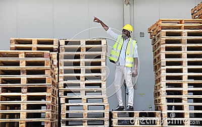 African American worker in safety vest suit and hardhat standing on the pile of pallet wood at warehouse Stock Photo