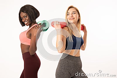 African and european sporty girls in sportswear standing back to back, international girls holding yoga mats on their Stock Photo
