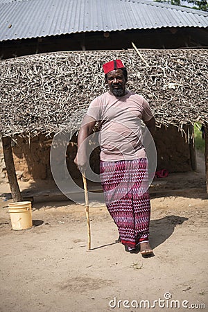 An African Older Man in Red Muslim Taqiyyah Fez Hat posing with a stick for lame people on Yard Near the Basic Hut with Stock Photo