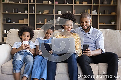 African couple with kids sit on sofa with diverse gadgets Stock Photo