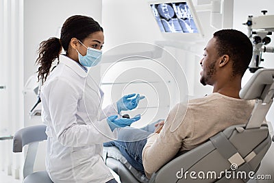 African Stomatologist Woman Discussing Teeth Treatment Process With Male Patient In Clinic Stock Photo