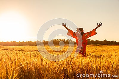 African woman in traditional clothes standing arms raised in a field of crops at sunset or sunrise Stock Photo