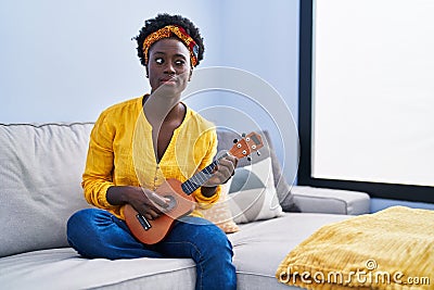 African young woman playing ukulele at home winking looking at the camera with sexy expression, cheerful and happy face Stock Photo