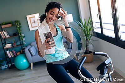 Afro young fitness girl using mobile phone while training on exercise bike at home Stock Photo