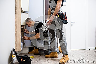 Aged electrician, repairman in uniform working, examining ethernet cable or router in fuse box using flashlight while Stock Photo