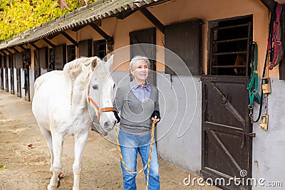 Aged farmer woman leading white horse outdoors along stables Stock Photo