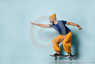 Aged male in t-shirt, orange pants, hat, gumshoes. Riding black skateboard, clenched fists, posing sideways on blue background Stock Photo