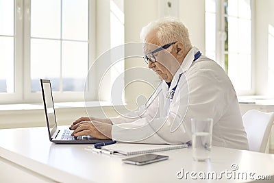 Aged professional doctor in uniform sitting at laptop and communicating with patient online Stock Photo