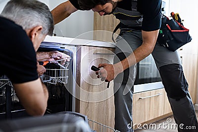 Aged repairman fixing dishwasher in the kitchen, using screwdriver while his young colleague in uniform helping him Stock Photo