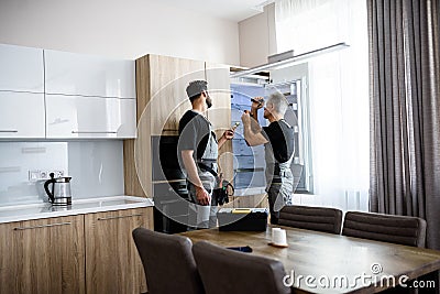 Aged repairman in uniform fixing refrigerator in the kitchen, while his colleague helping him, bringing screwdriver Stock Photo