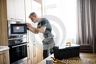 Aged repairman in uniform working, examining broken microwave in the kitchen using flashlight. Repair service concept Stock Photo