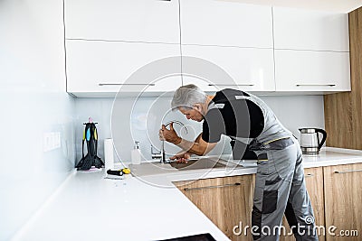 Aged repairman in uniform working, fixing broken kitchen tap using adjustable wrench. Repair service concept Stock Photo