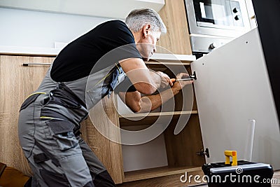 Aged repairman in uniform working, fixing kitchen cabinet using screwdriver. Repair service concept Stock Photo