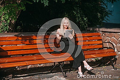 aged woman with long blond hair, in a black long dress, remotely works on a tablet, sits on a bench Stock Photo