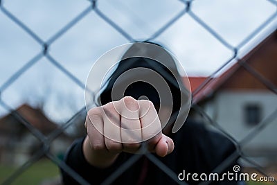 Aggressive teenage boy showing hes fist behind wired fence at the correctional institute, focus on the boys hand Stock Photo