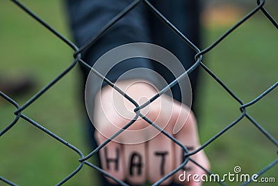 Aggressive teenage boy showing hes fist behind wired fence at the correctional institute, the word hate is written on hes hand Stock Photo