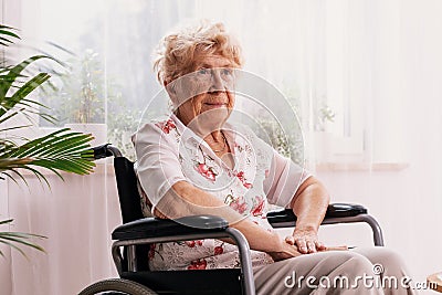 Ailing woman dressed in a blouse with flower pattern is sitting in a wheelchair in her home Stock Photo