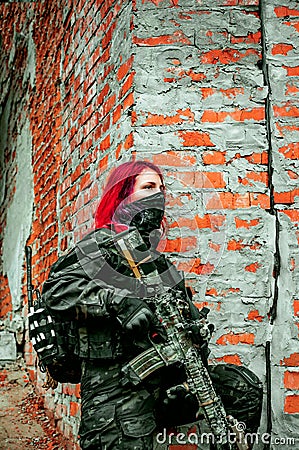 Airsoft red-hair woman in uniform with machine gun standing beside brick wall. Soldier on ruine. Vertical photo Stock Photo