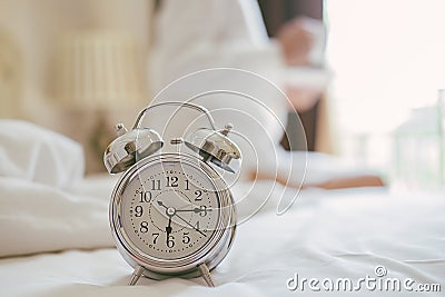 Alarm clock standing on bedside table has already rung early morning to wake up woman is stretching in bed in Stock Photo