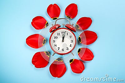 Alarm clock with tulip petals around. Flat lay style, over blue background. Daylight savings time concept. Spring Forward. Stylish Stock Photo