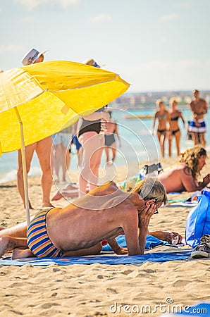 Alcudia, Spain 14.09.2011 - Older man reading a newspaper at sandy beach under umbrella. People sunbathing at Playa de Muro. Editorial Stock Photo