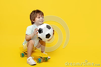 Amazed little boy play with soccer ball while sitting on pennyboard or skateboard over yellow background. Stock Photo