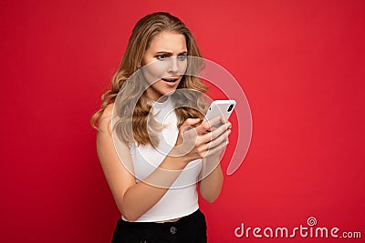 Amazed shocked beautiful young blonde woman wearing white t-shirt isolated over red background using smartphone and Stock Photo