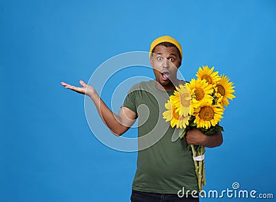 Amazed, shocked young African American man with armful of sunflowers pointing with hand up wearing green t-shirt and yellow hat Stock Photo