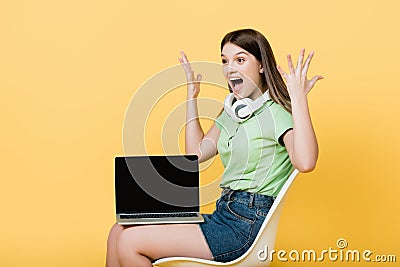 Amazed teenager with headphones and laptop sitting on chair isolated on yellow. Stock Photo