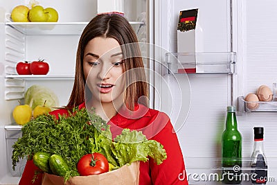 Amazed young female with surprised expression looks at vegetables, forgets to buy something in grocer`s shop, stands in kitchen ne Stock Photo