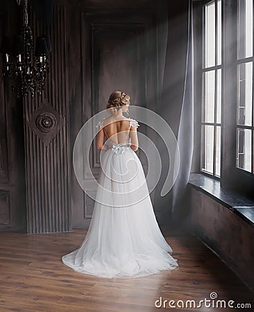 Amazing lady in long white adorable expensive light dress with train and open back stands with back to camera, girl with Stock Photo