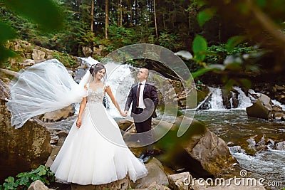 Amazing romantic view of happy bride with groom near beautiful grand waterfall in mountain. luxurious wedding dress. Marriage coup Stock Photo