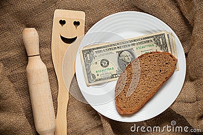 American dollar and a piece of black bread on a white plate and next to it lies a kitchen wooden spatula with a smiley face and a Stock Photo