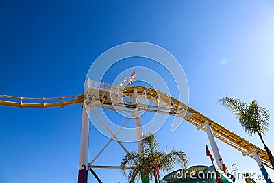 An American flag and a California flag on top of a yellow rollercoaster with a gorgeous blue sky and tall lush green palm trees Editorial Stock Photo