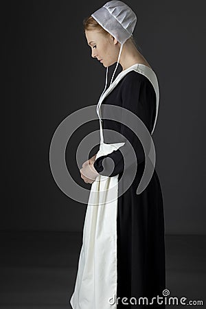 An Amish woman wearing a long black dress, apron, and cap against a studio backdrop Stock Photo