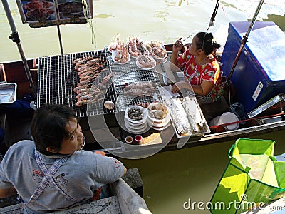 A woman cooks barbecue seafood in a boat while the waitress waits for the order at Amphawa Floating Market Editorial Stock Photo