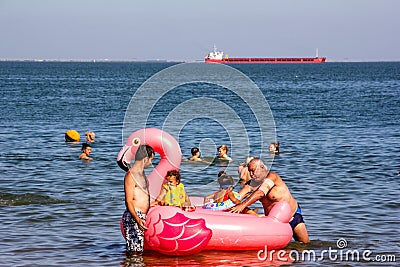 Young and old men ride small children on an inflatable pink flamingo and other vacationers and tourists swim in the sea Editorial Stock Photo