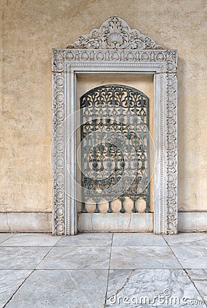 Ancient ornate green iron grid framed by ornate marble frame over beige stone wall and white marble tiled floor Stock Photo