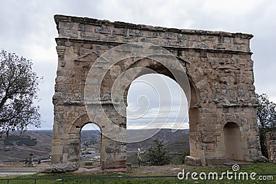 An ancient, weathered stone arch with two openings stands against a backdrop of a cloudy sky and distant landscape Stock Photo