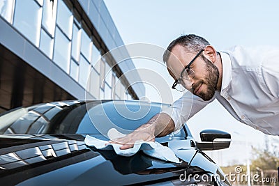 Angle view of happy businessman in glasses cleaning black car with white cloth near building Stock Photo