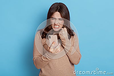 Angry casual woman using smart phone isolated on blue background, frowning face and clench fist with anger, looks at display, Stock Photo