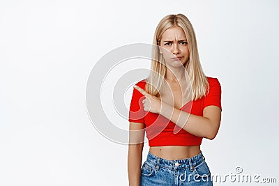 Angry sulking woman pointing finger left and frowning, upset about something, complaining, standing against white Stock Photo