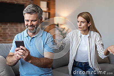 Angry upset adult european wife yelling at smiling husband with smartphone in living room interior Stock Photo